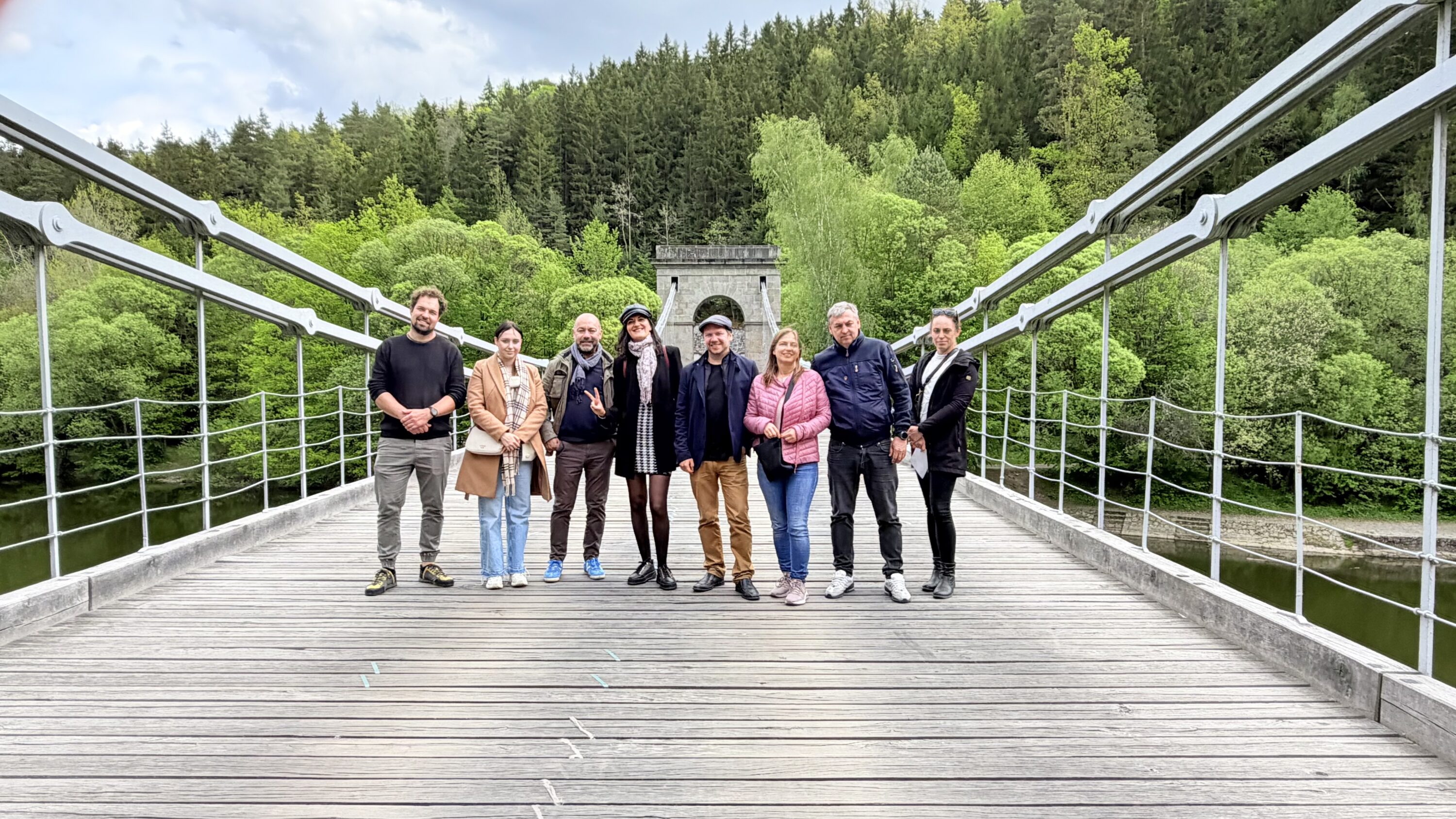 Nine adults pose together on a wooden suspension bridge, with a lush green forest behind them.