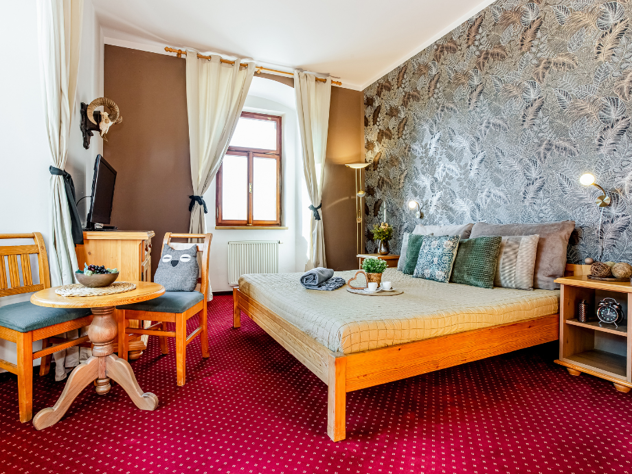 Cozy hotel room with a wooden bed, patterned wallpaper, and a red carpet, plus a small dining table with two chairs nearby.