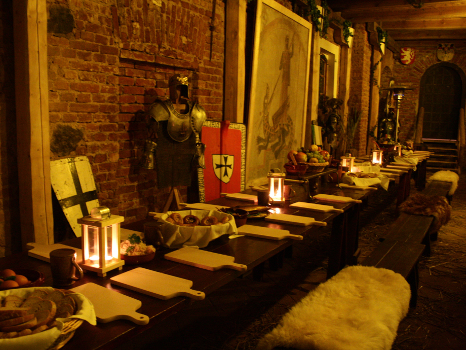 Long medieval banquet table in a brick-walled hall, set with cutting boards, lanterns, armor, shields, and bread and fruit.