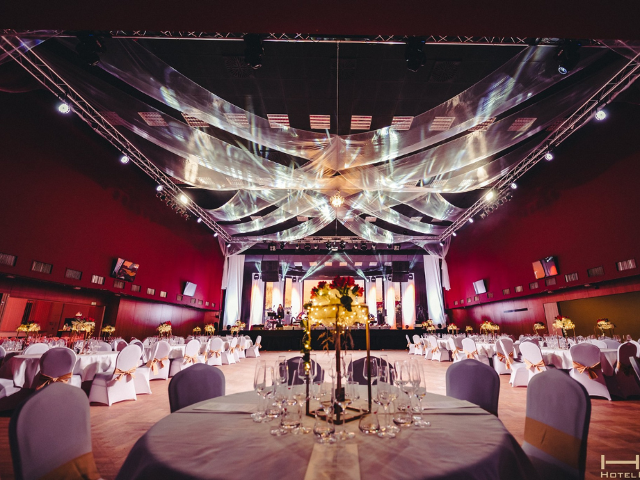 Elegant banquet hall set for a formal reception: round tables with white chair covers, gold centerpieces, and a stage with lighting at the far end under a draped ceiling.