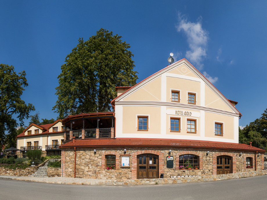 Hotel Gold: a two-story beige building with a stone ground floor, red roof, and a blue sky behind, surrounded by trees.