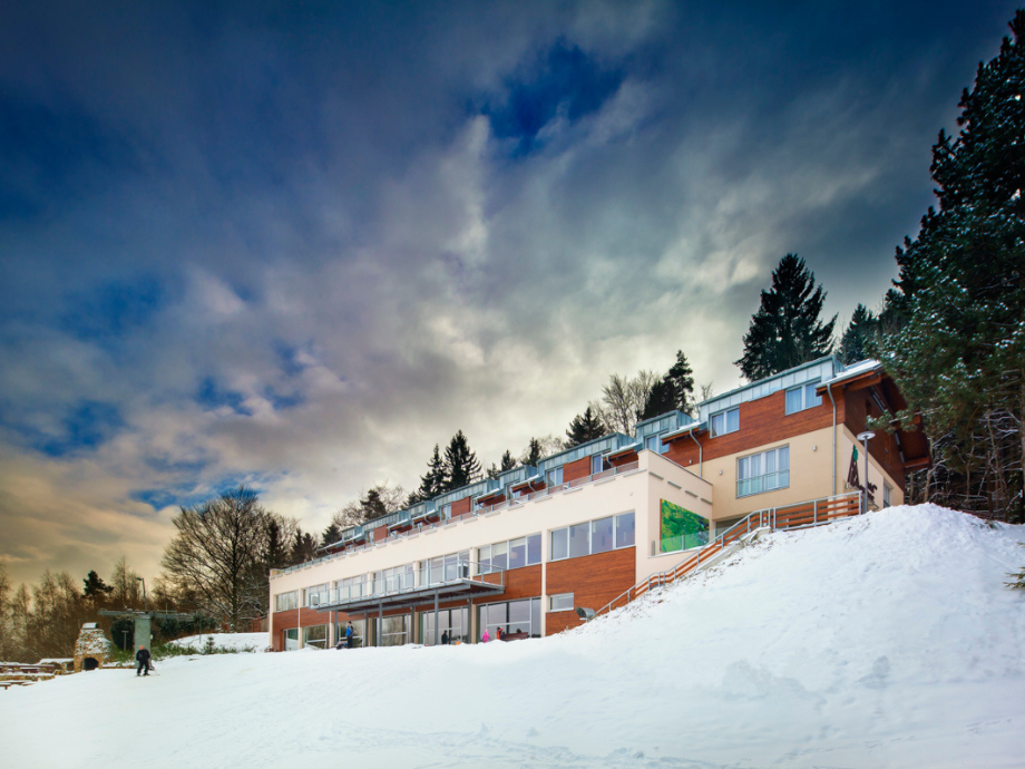 Modern snow-covered lodge/hotel with large windows on a hillside and evergreen trees under a dramatic sky.