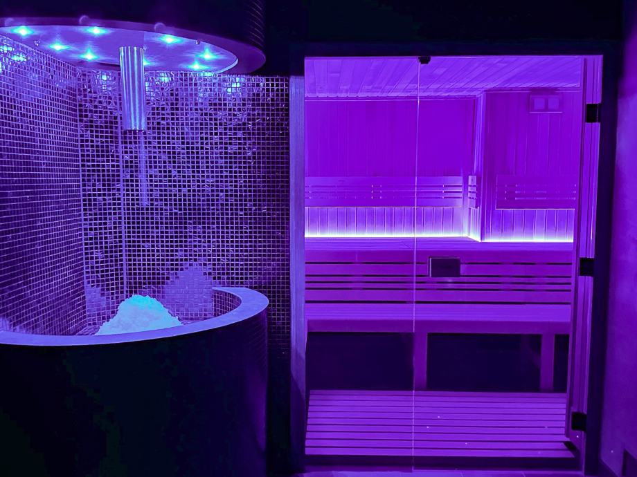 Purple-lit spa sauna interior with a glass door, tiled wall on the left, and a wooden bench on the right; a bucket of ice sits near the shower area.