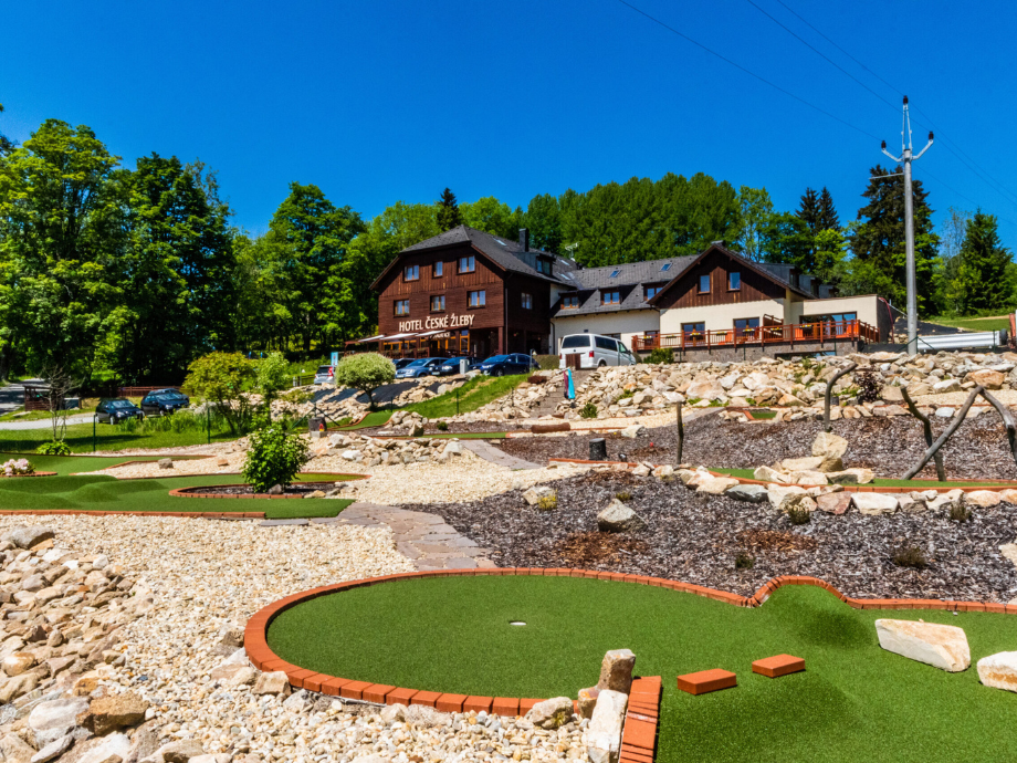 Wooden hotel building with sign 'Hotel Ceske Zleby' atop a rocky landscape, overlooking a mini‑golf course on a sunny day.
