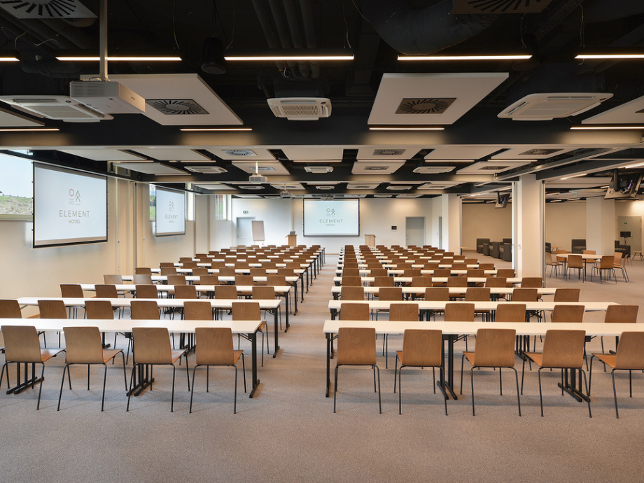 Large conference room with rows of wooden chairs and white tables facing a screen displaying Element Hotel.