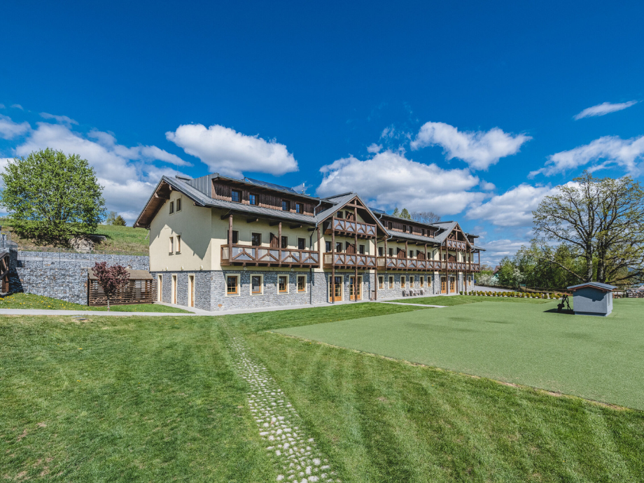Chalet-style hotel with wooden balconies and a stone base, set on a manicured lawn under a bright blue sky.