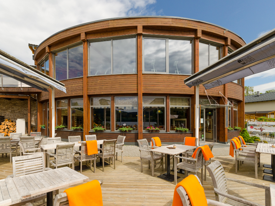Outdoor cafe seating in front of a curved wooden building with large windows and orange-cushioned chairs.