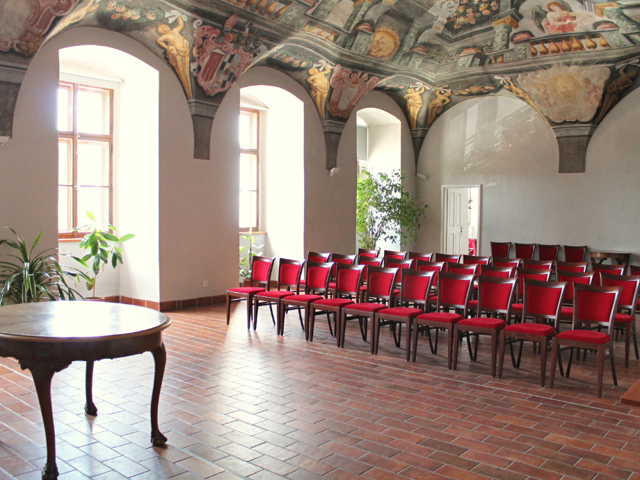 Historic hall with ornate vaulted, painted ceilings, red-upholstered chairs in rows, and a round wooden table in the foreground on a brick floor.
