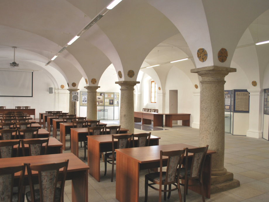 Study hall or classroom with stone arches and columns, rows of wooden tables and chairs under arched ceiling.