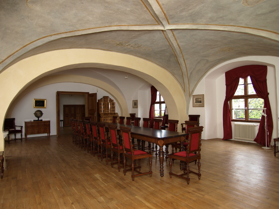 Formal meeting room with a long wooden table and many red-upholstered chairs under vaulted ceilings with arched alcoves and windows with red drapes.