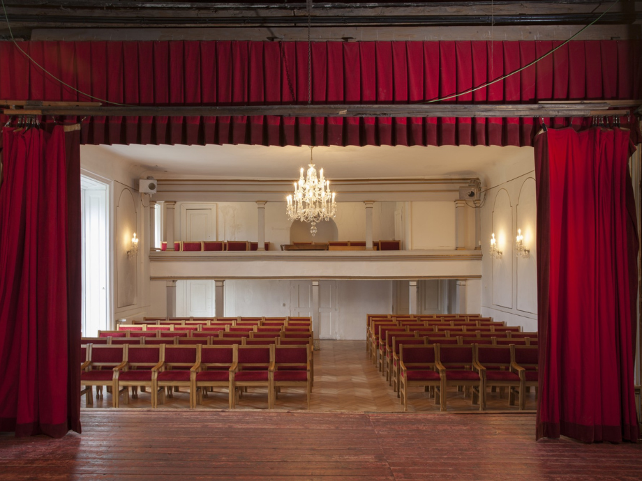 Empty theater auditorium viewed from backstage, red upholstered seats, chandelier, and balconies.