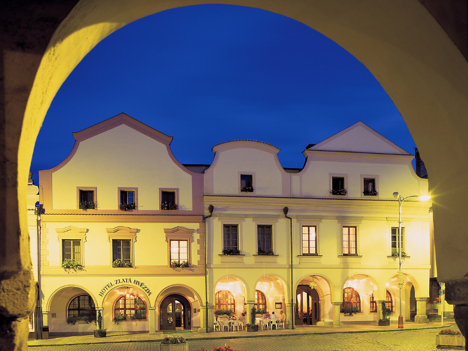 Nighttime view of a pastel-yellow, historic hotel with arched colonnade and flower boxes, framed by a stone archway outside the building.