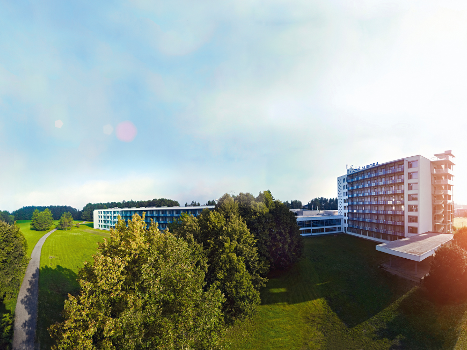 Aerial view of a modern hotel complex surrounded by green lawns and trees, with a winding path on the left.