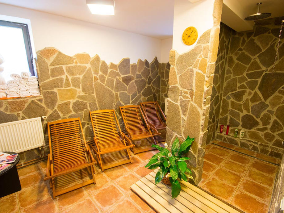 Stone-walled spa relaxation area with five wooden lounge chairs, towels by the window, a yellow wall clock, and a plant near a tiled shower space.