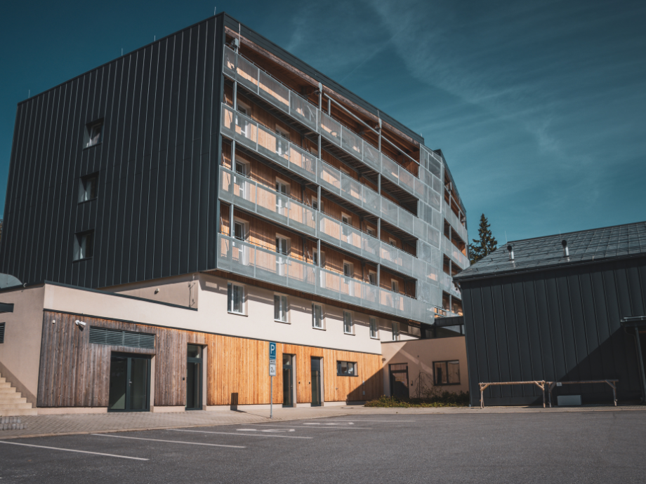 Modern six-story building with glass balconies and a wood-clad ground floor beside a parking lot under a blue sky.