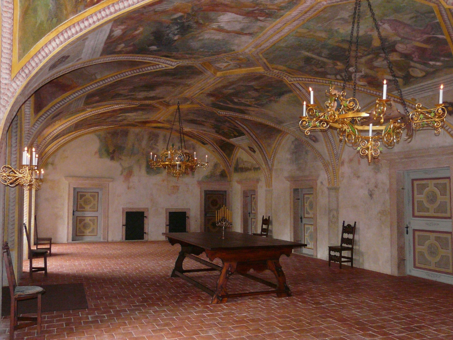Historic vaulted room with painted arched ceiling, ornate chandeliers, and a large wooden table with chairs.