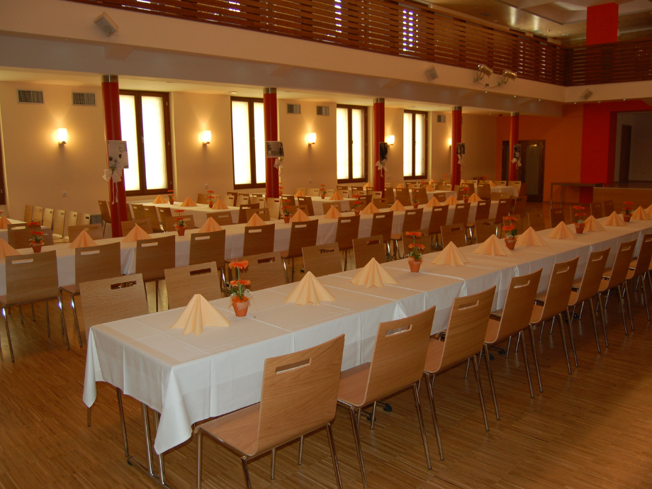 Banquet hall prepared for a formal event with long white-tablecloth tables, folded napkins, and small potted flowers, surrounded by wooden chairs.