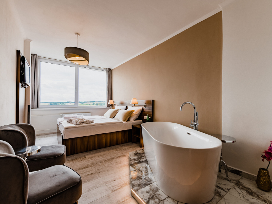 Hotel room with a freestanding oval bathtub in the foreground, a bed and seating area by a large window overlooking the countryside.