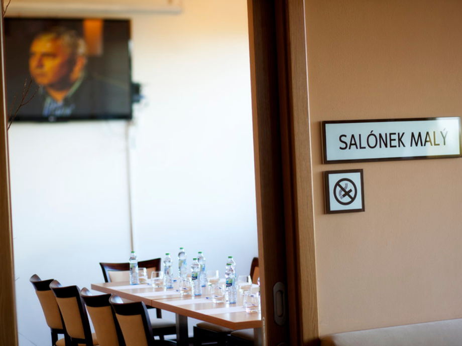 Conference room seen through an open door: table set with water bottles and glasses, with a blurred TV showing a man in the background.