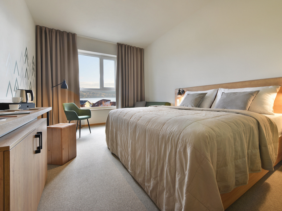 Hotel room with a large bed, beige comforter, two pillows, and wooden headboard; desk and green chair by a window with brown curtains.