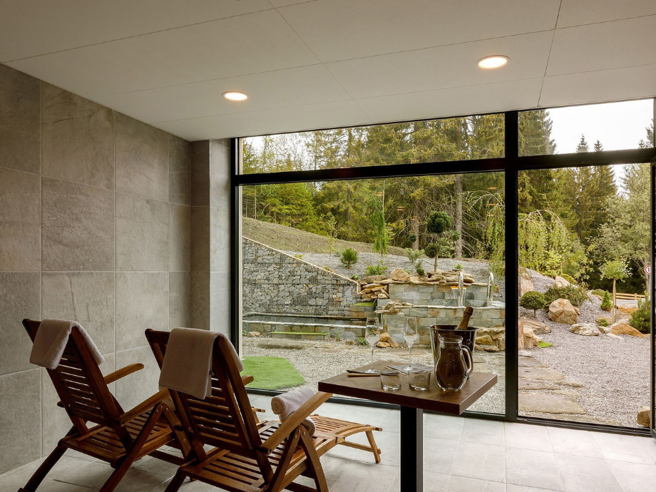 Indoor spa/wellness area with two wooden lounge chairs facing a large glass wall, overlooking a stone garden and greenery outside. A small table holds a pitcher and glasses.