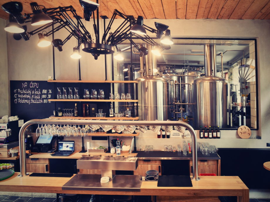 Industrial bar counter with glassware, taps, and large stainless steel brewing tanks visible through a pass-through window.