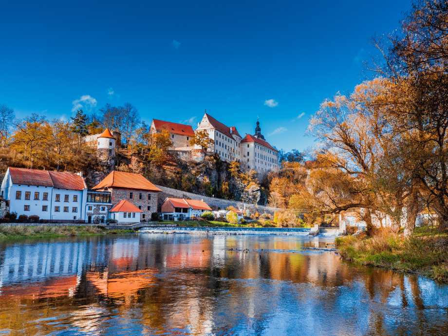 Autumn riverside scene with a castle on a hill, colorful trees, and reflections in the water