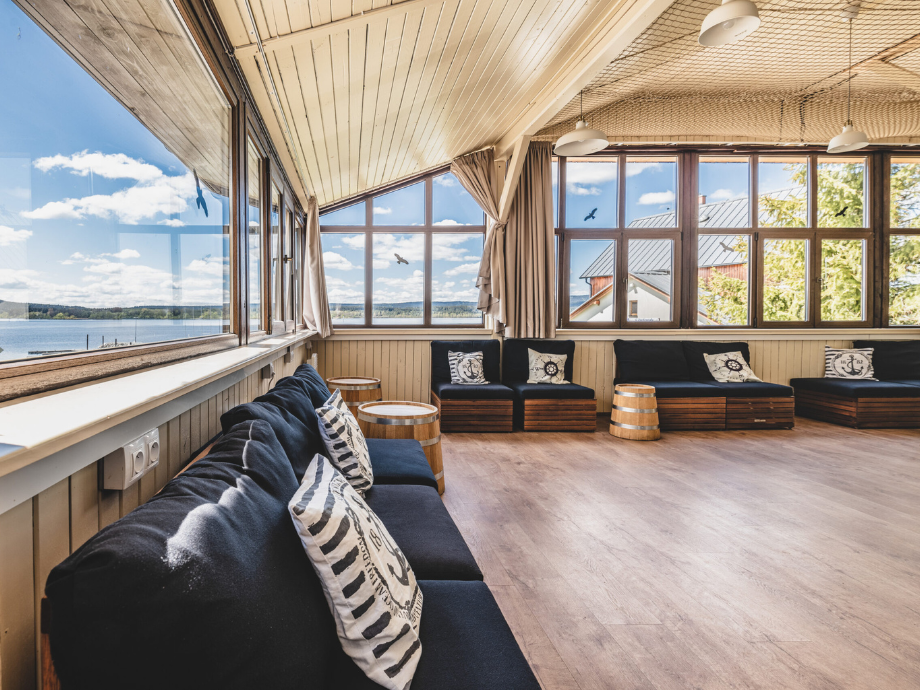 Sunlit lakeside lounge with dark blue couches, nautical cushions, and large window walls showing a blue lake outside.