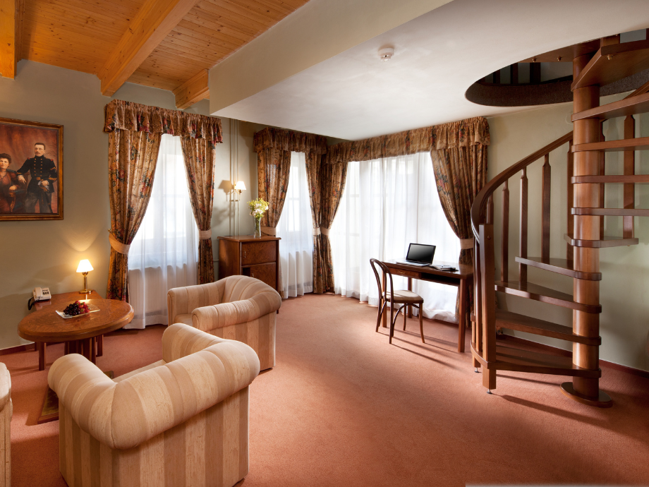 Cozy hotel-style living room with two beige striped armchairs, a round coffee table, warm lamps, and heavy floral curtains; a wooden spiral staircase rises along the wall. A desk with a laptop sits by the windows, creating a small workspace.
