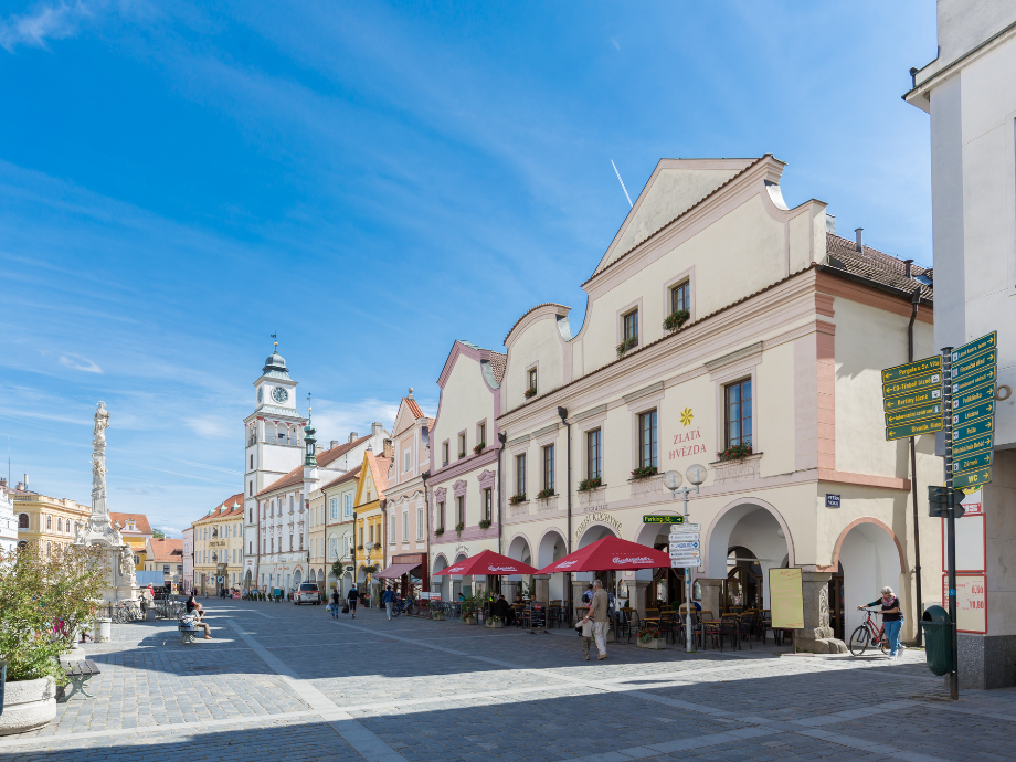 Pastel-colored town square with outdoor cafe tables under red umbrellas and a clock tower in the background. Attractively lit on a sunny day.