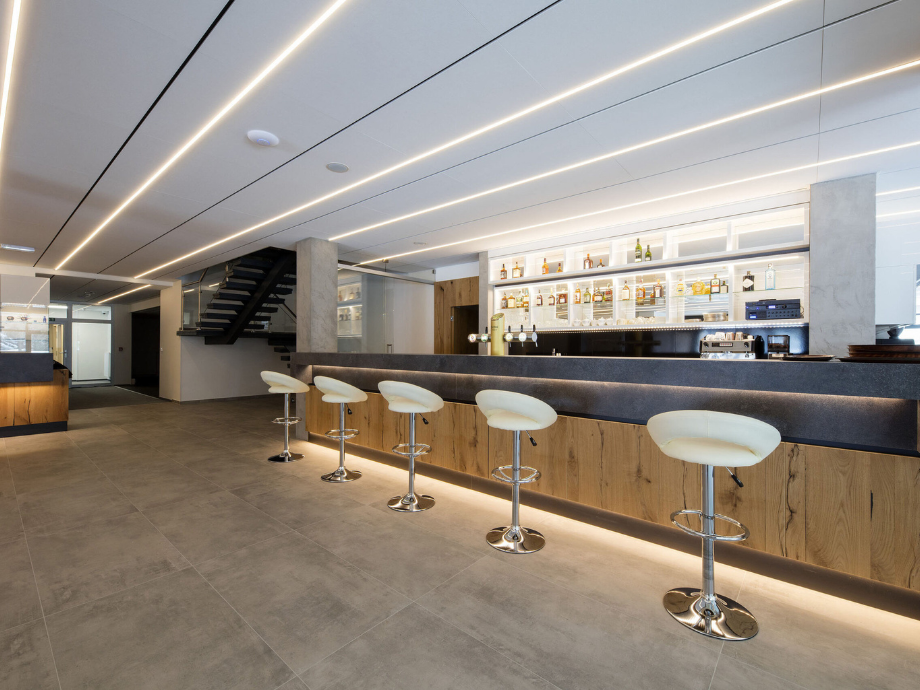 Modern bar with white stools along a wooden and dark countertop, backlit bottles on illuminated shelves behind the counter.