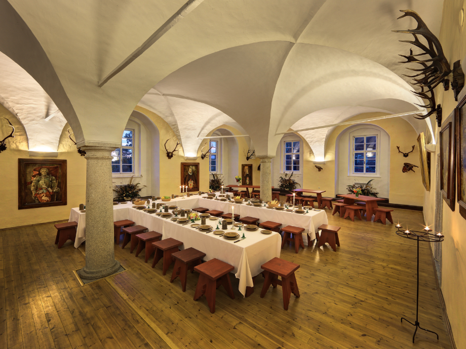Historic dining hall with vaulted white arches, stone columns, and long banquet tables set with place settings and candles.