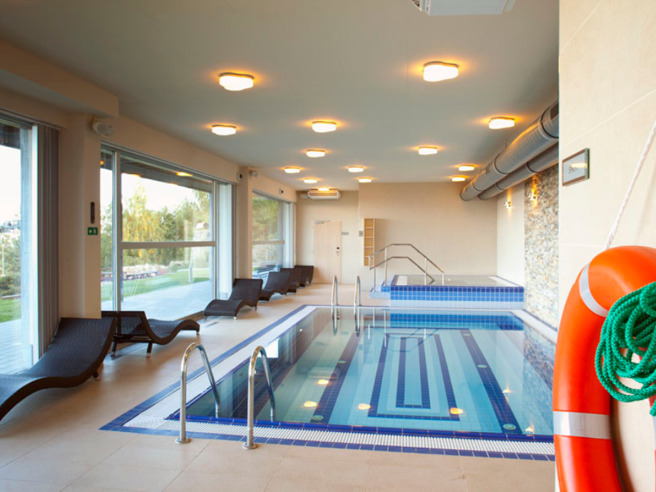 Indoor hotel-style swimming pool with blue tiled lanes, ladders, and a row of lounge chairs along the left; large windows outside; orange lifebuoy with rope on the right.