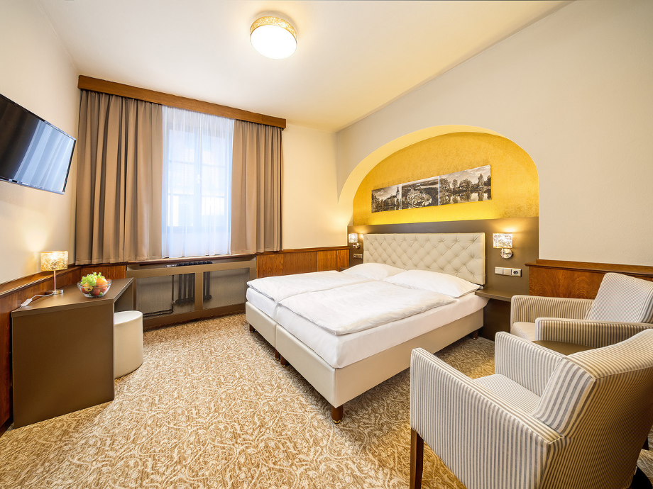 Hotel room with a large bed and tufted headboard under a yellow arched wall, two striped armchairs, and a window with curtains on the left side.
