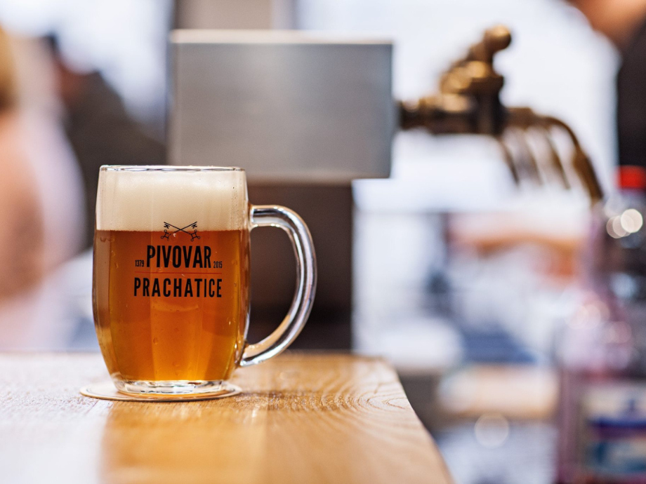 Beer mug with a foamy head on a wooden bar; logo reads PIVOVAR PRACHATICE in bold black letters on the glass surface.