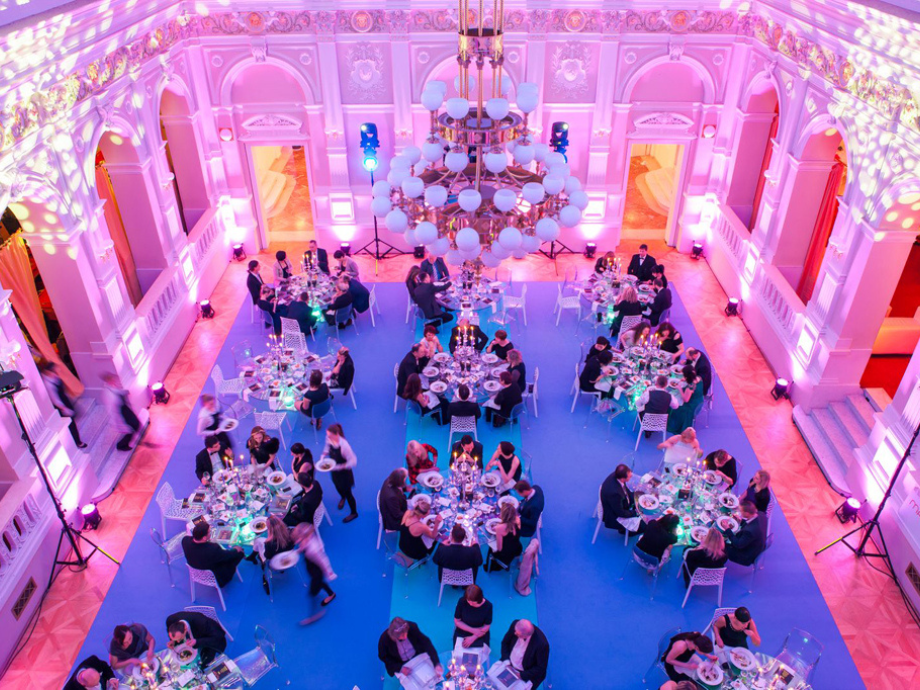 Guests seated at round tables for a formal gala in a grand ballroom with pink-purple lighting and a large chandelier above.