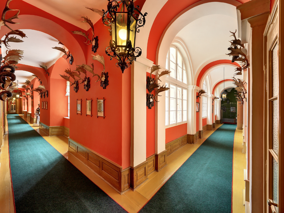 Long, red-walled corridor with white arches, hanging lanterns, and mounted deer antlers along the walls in a historic building.