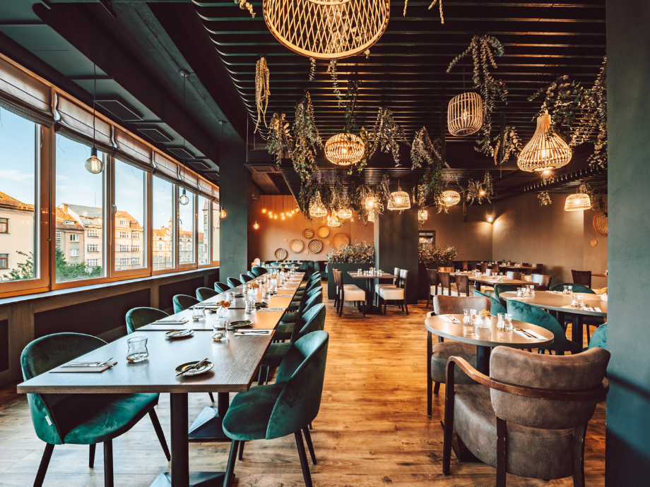 A warmly lit restaurant with a long communal table along windows and green upholstered chairs, wicker pendant lights overhead.