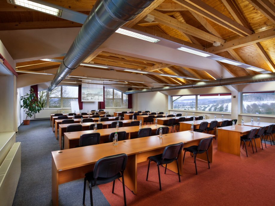 Lecture hall with rows of wooden desks and black chairs, water bottles at each seat, and large windows along the wall.