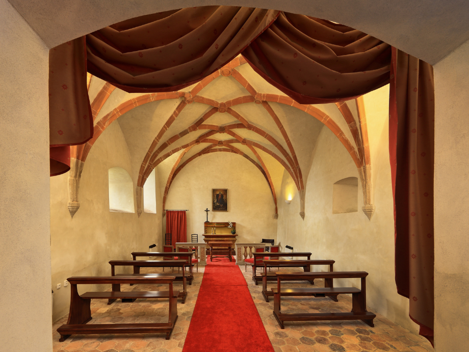 Interior of a small chapel with a red carpet aisle, wooden pews, and Gothic vaulted arches leading to an altar area.