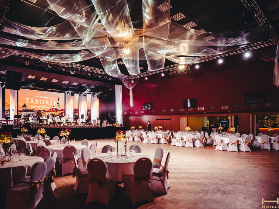 Banquet hall with round tables dressed in white, flower centerpieces, and a live band on a lit stage in the background.