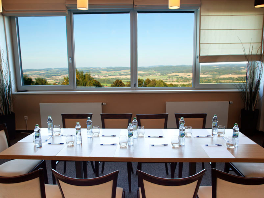 Meeting room with a long table set for eight, water bottles, glasses, pens, and notepads, view of countryside through large windows.
