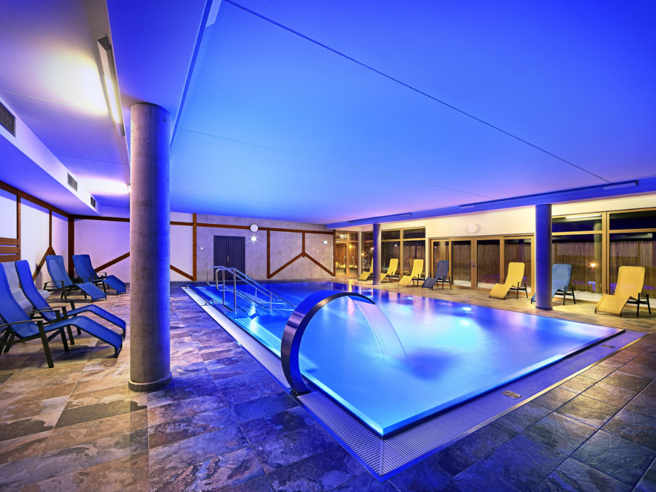 Indoor pool area with blue lighting, a curved water slide, and rows of lounge chairs along the walls.