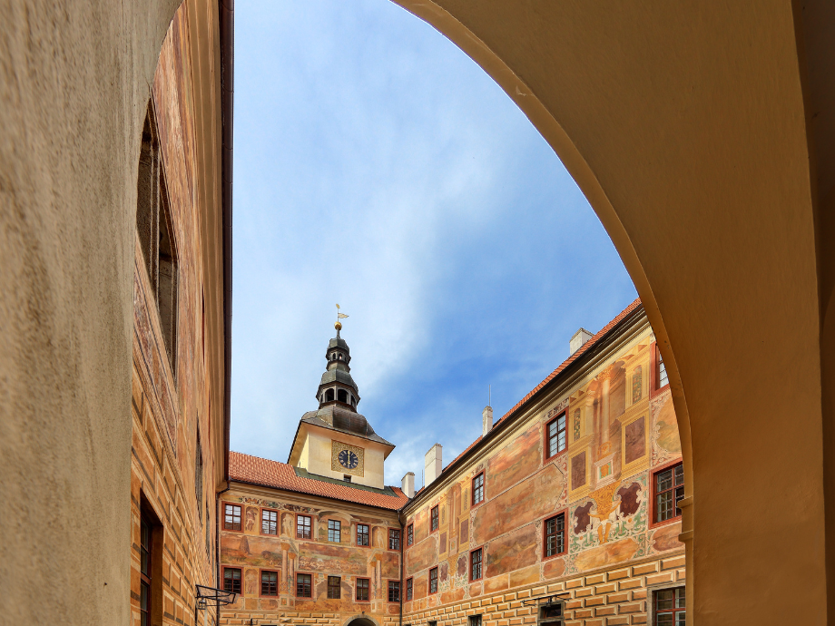 Inner courtyard of a historic building with frescoed walls, a clock tower, and a blue sky framed by an arch/arcade window.