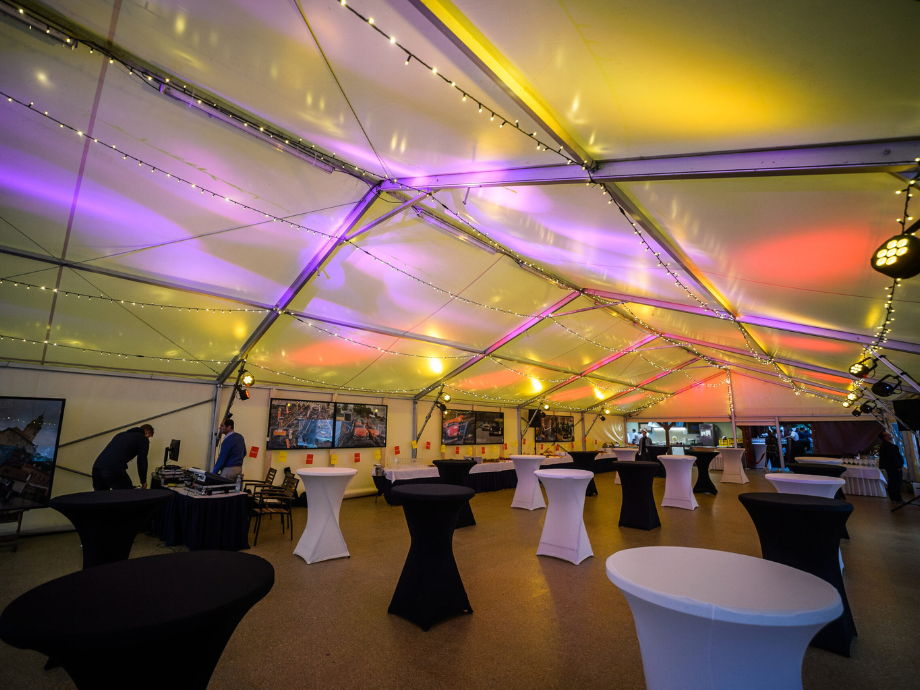 Event tent interior with string lights and colorful stage lighting, set for a cocktail reception with tall tables covered in black and white cloths far apart from a DJ area on the left.