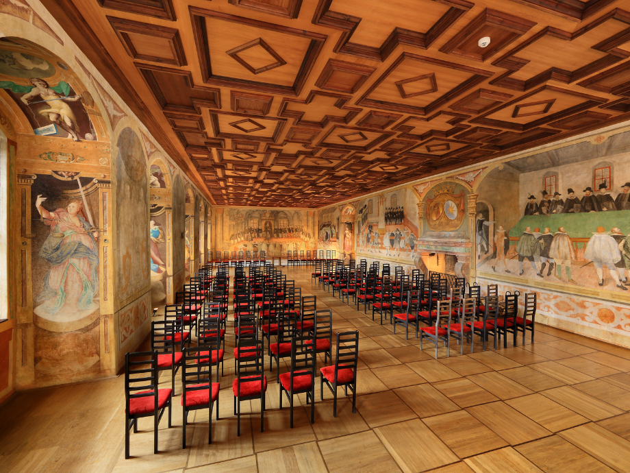 Frescoed banquet hall with a coffered wooden ceiling and rows of black chairs with red cushions facing forward, a ceremonial room ready for an event.