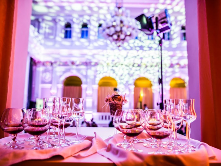 Banquet table with many wine glasses and a small centerpiece in a purple-lit grand hall.
