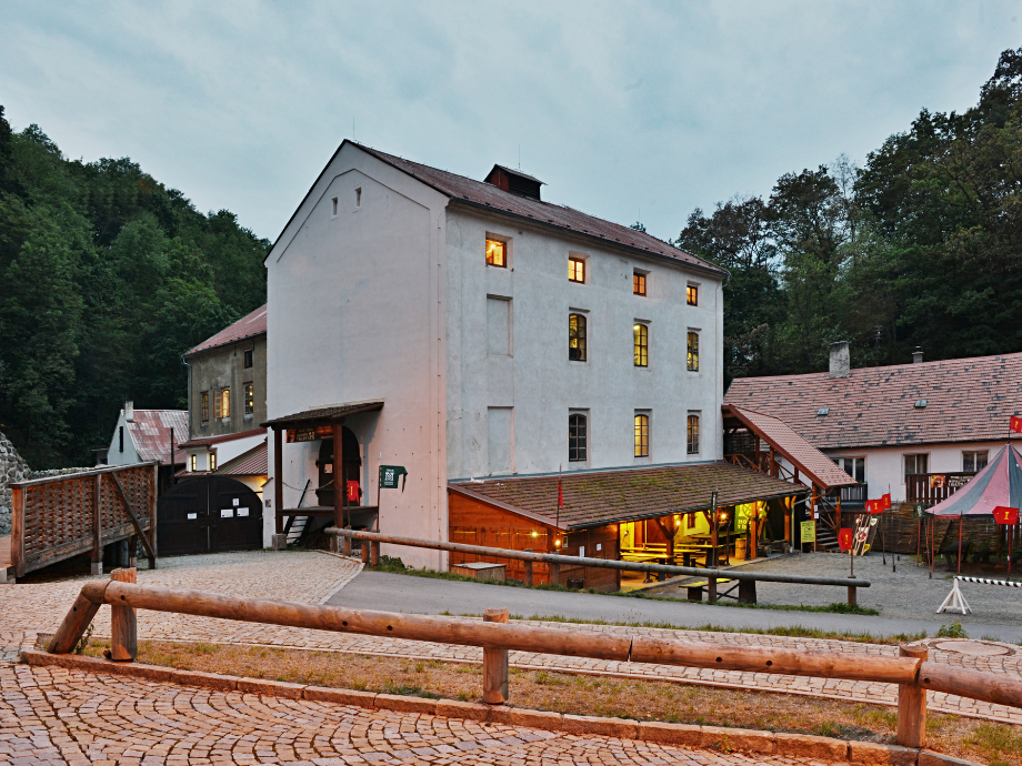 White three-story building with warm-lit windows in a forested setting; cobblestone courtyard and wooden railing in the foreground.