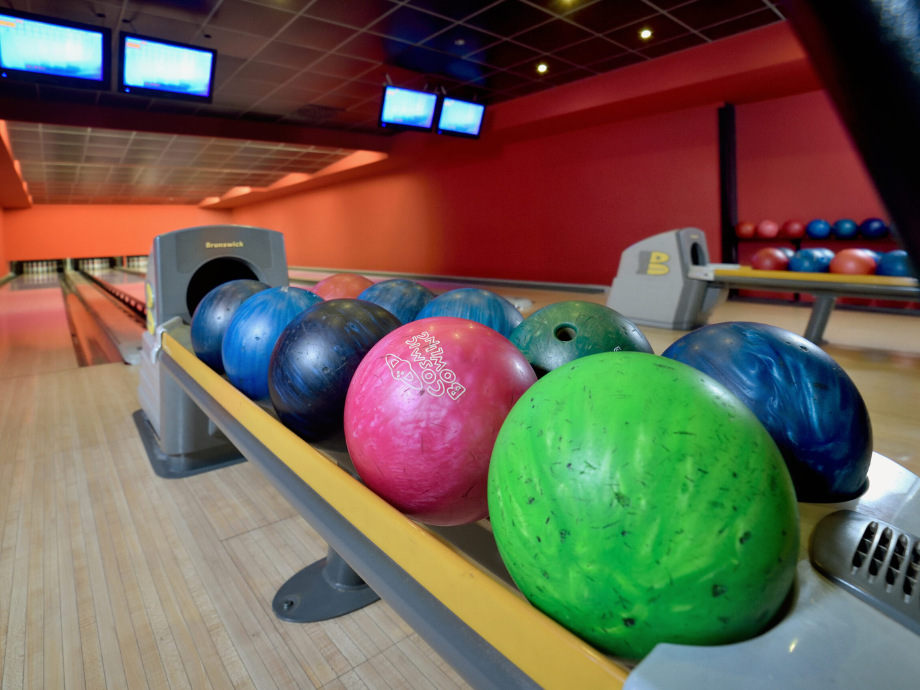 Bowling balls lined up on a rack at a bowling alley, with lanes and overhead score monitors in the background.