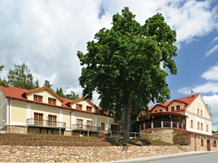Large leafy tree in front of a yellow, two-story building with red tiled roof and stone retaining wall under a blue sky with clouds